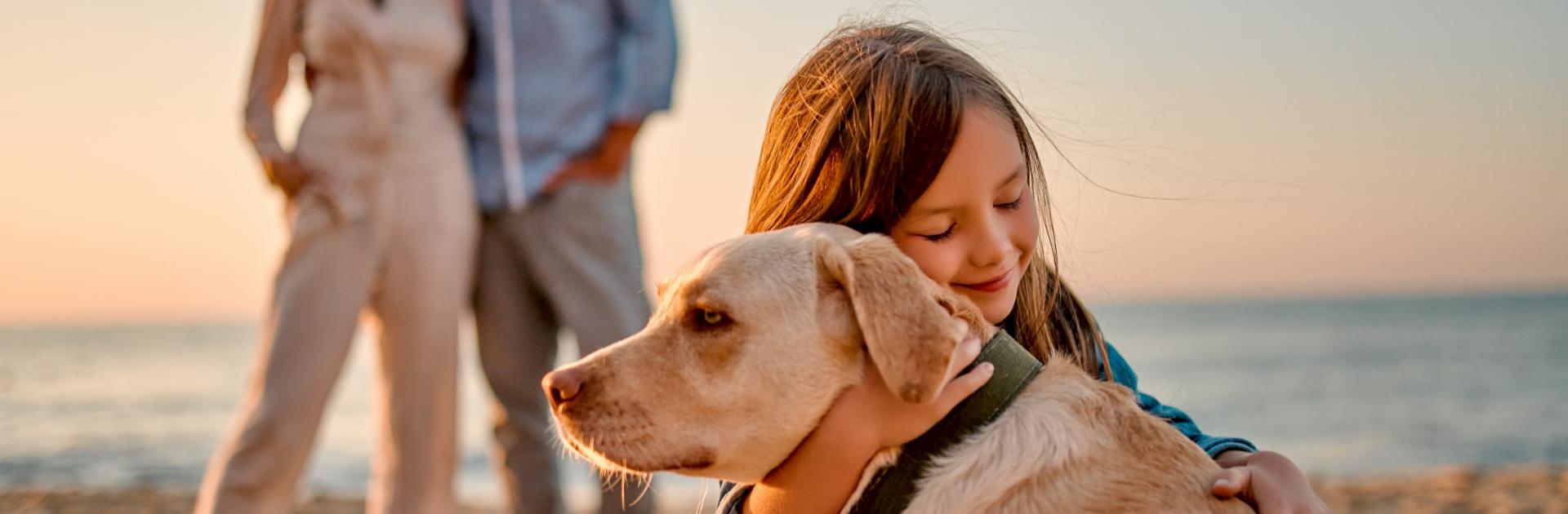 Bambina abbraccia un cane sulla spiaggia, con i genitori sullo sfondo.