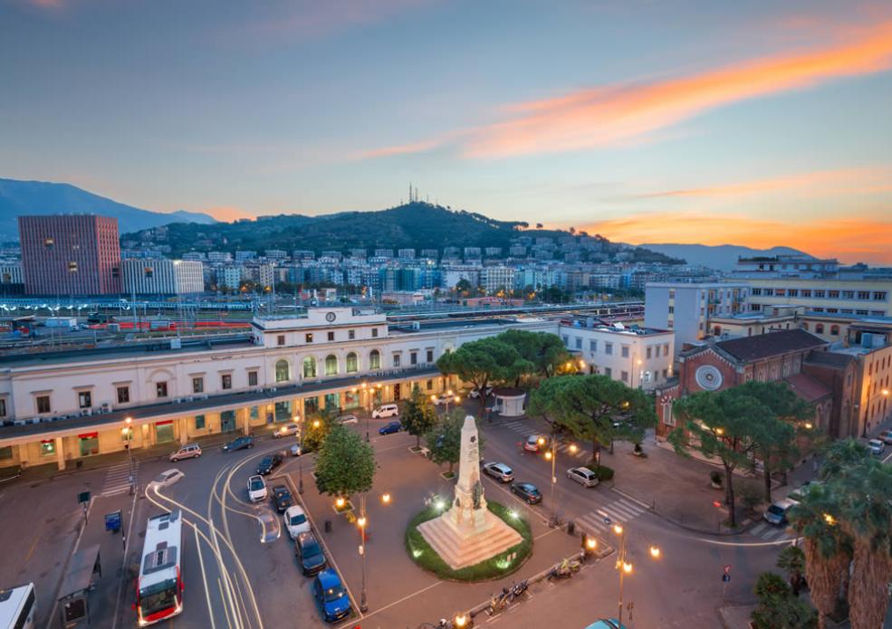 Veduta serale di una piazza con monumento e stazione ferroviaria sullo sfondo.